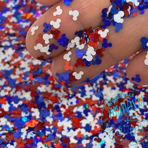 May include: Red, white, and blue glitter in the shape of Mickey Mouse heads. The glitter is scattered on a surface and a hand is holding some of the glitter.