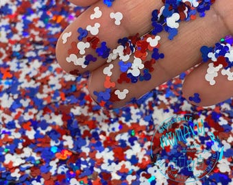 May include: Red, white, and blue glitter in the shape of Mickey Mouse heads. The glitter is scattered on a surface and a hand is holding some of the glitter.