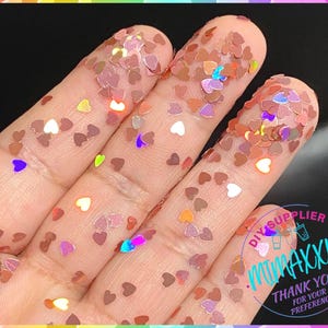 May include: A close-up of a hand covered in iridescent pink heart-shaped glitter. The glitter is scattered across the hand and fingers, creating a sparkly and festive look.