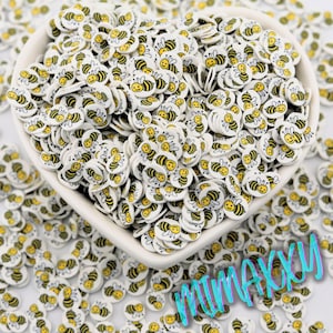 May include: A heart-shaped bowl filled with small, white, circular embellishments. Each embellishment features a smiling yellow and black bee design. The background is scattered with similar bee-themed embellishments. The word "MIMAXXY" is visible in the bottom right corner.