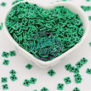 May include: A white heart-shaped bowl filled with green and black Minecraft character sprinkles. The sprinkles are small and square-shaped with a black face on each one.