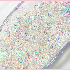 May include: A close-up of iridescent glitter in a clear container. The glitter is a mix of white, silver, and rainbow colors.