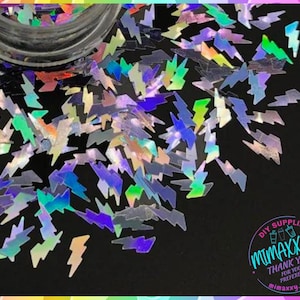 May include: A close-up of iridescent holographic glitter in the shape of lightning bolts. The glitter is scattered on a black surface. The glitter is perfect for adding a touch of sparkle to crafts, nail art, and resin projects.