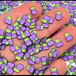 May include: Purple and green slime charms with a monster face design. The charms are small and perfect for adding to slime, resin, or other crafts.