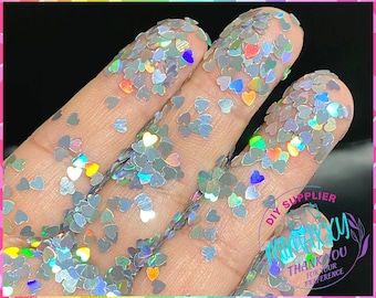 May include: Silver iridescent heart-shaped glitter. The glitter is scattered on a hand, showing the size and shape of the glitter.
