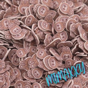 May include: A pile of small, brown and white polymer clay pieces shaped like a cartoon sloth. The sloth faces have a smiling expression and are surrounded by a white border. The image has a close-up view.