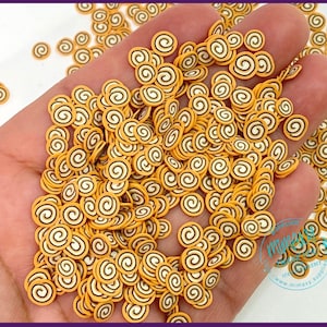 May include: A hand holding a large quantity of small, yellow and brown swirl-shaped polymer clay sprinkles. The sprinkles are perfect for adding a decorative touch to crafts, resin projects, and other DIY projects.