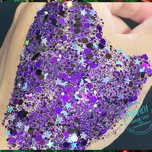 May include: Purple, black, and iridescent glitter with star and crescent moon shapes. The glitter is on a hand, and the background is a green plant.