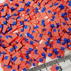 May include: A large pile of small red, white, and blue American flag-themed polymer clay charms. The charms are approximately 1 cm in size and are scattered on a white surface. A ruler is visible in the bottom right corner of the image.