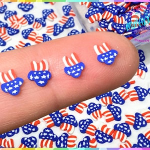 May include: A close-up of a fingertip with several small, red, white, and blue American flag-themed charms. The charms are shaped like a heart with a star in the center. The charms are scattered on a white surface.