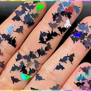 May include: A close-up of a hand holding a variety of iridescent black cat-shaped glitter. The glitter is scattered across the hand and fingers, creating a sparkly and festive look.