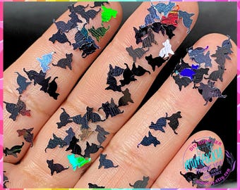 Op de afbeelding: Een close-up van een hand die een verscheidenheid aan iriserende zwarte kattenvormige glitter vasthoudt. De glitter is verspreid over de hand en vingers, waardoor een sprankelende en feestelijke look ontstaat.