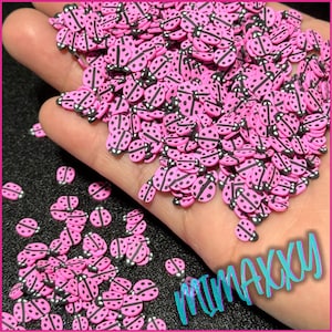 May include: Pink and black ladybug shaped sprinkles. The sprinkles are scattered on a hand and a black surface.