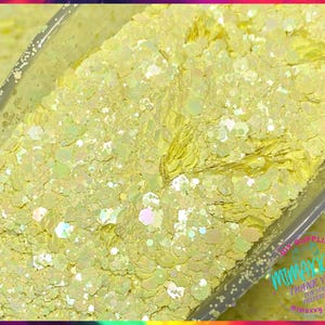 May include: Close-up of a container filled with pale yellow glitter. The glitter has a mix of shapes and sizes, including hexagonal and irregular pieces. Some pieces have an iridescent sheen, reflecting multiple colors. The container has text that reads "DIY SUPPLIER MIMAXXY".