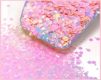 May include: Pink, iridescent glitter with a variety of hexagon shapes. The glitter is in a clear plastic container with the text "SUPPLIER" on the side.