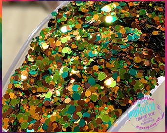 May include: A close-up of a container filled with green, gold, and brown glitter. The glitter is iridescent and has a variety of shapes and sizes.