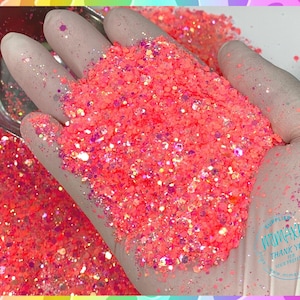 May include: A close-up of a hand holding a large amount of pink glitter with iridescent accents. The glitter is in a bowl and is spilling out onto a surface.