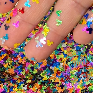 May include: A close-up of a variety of colorful iridescent glitter shaped like Mickey Mouse heads. The glitter is scattered on a surface and a hand is holding some of the glitter.