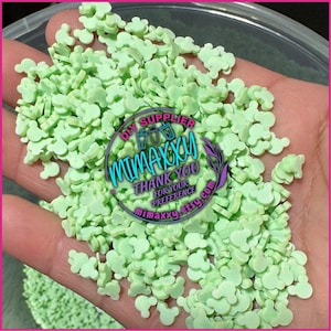 May include: A close-up of a hand holding a large amount of light green Mickey Mouse head shaped sprinkles. The sprinkles are in a bowl and the text "DIY SUPPLIER MIMAXXY THANK YOU FOR YOUR PREFERENCE MIMAXXY.ETSY.COM" is visible on a sticker.