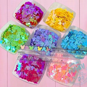 May include: Six containers of iridescent glitter shaped like Mickey Mouse heads. The glitter is in various colors, including pink, green, blue, yellow, purple, and orange. A ruler is in the image for scale.
