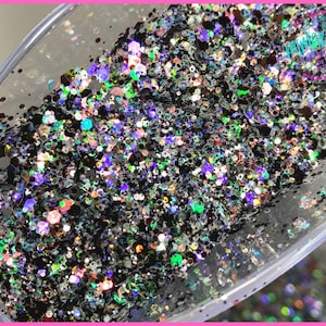 May include: A close-up of a scoop filled with black glitter with iridescent and holographic accents. The glitter is a mix of different sizes and shapes.