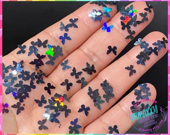 May include: A close-up image of iridescent black butterfly shaped glitter. The glitter is scattered on a hand, with some of the glitter pieces overlapping.