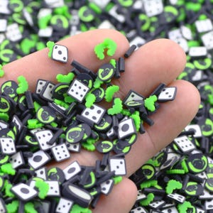 May include: A close-up of a hand holding a variety of black and green resin dice and other shapes. The dice are white with black dots and the other shapes are black with green details.
