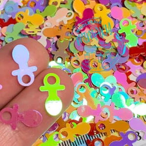 May include: A close-up of a variety of iridescent baby pacifier shaped confetti in various colors, including pink, blue, green, yellow, and orange. The confetti is scattered on a surface and a hand is holding some of the confetti.