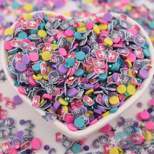May include: A heart-shaped bowl filled with a colorful assortment of small, flat, circular clay slices featuring cartoon character faces, alongside solid yellow, pink, purple, and teal circles, and small iridescent rhinestones.