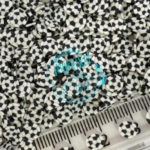 May include: Black and white soccer ball sprinkles. The sprinkles are scattered on a white surface with a ruler in the background.