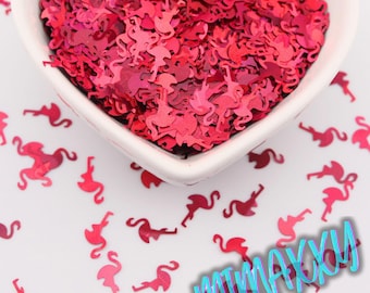 May include: A white heart-shaped bowl filled with red flamingo-shaped confetti. The confetti is scattered on a white surface, with some pieces outside the bowl. The confetti is a vibrant shade of red.