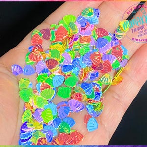 May include: A handful of iridescent holographic seashell shaped glitter. The glitter is in various colors including red, orange, yellow, green, blue, purple, and pink.