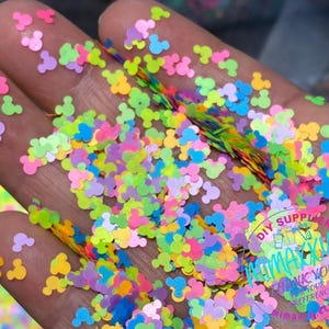 May include: A close-up of a hand holding a large amount of colorful Mickey Mouse head shaped glitter. The glitter is in a variety of colors, including pink, blue, green, yellow, and purple.