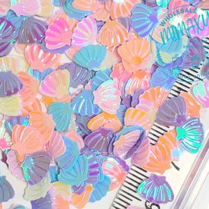May include: A collection of iridescent seashell-shaped sequins in various pastel colors, including pink, blue, purple, and yellow. The sequins are scattered on a white surface.