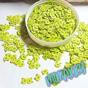 May include: A container filled with small, green, plastic, smiley face charms. The charms are scattered around the container.