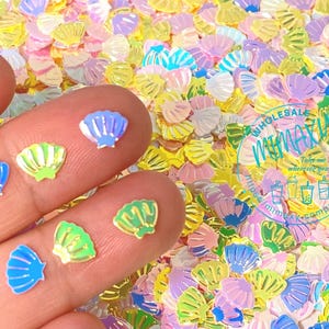 May include: Assortment of iridescent holographic seashell shaped glitter in various colors including pink, blue, green, and yellow. The glitter is scattered on a surface and a hand is holding some of the glitter.