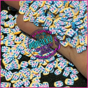 May include: A close-up of a variety of blue and white popcorn bucket sprinkles with the words "YUM", "MUY", and "MDY" printed on them. The sprinkles are scattered on a surface and a hand is holding some of the sprinkles.