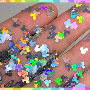 May include: A close-up of iridescent holographic Mickey Mouse head shaped glitter. The glitter is scattered on a hand, showing the different colors and shapes.