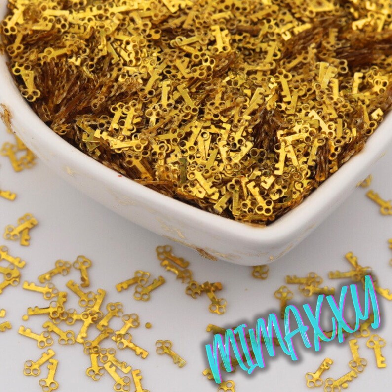 May include: A white bowl filled with small, gold-colored key charms. The charms are scattered around the bowl on a white surface. The image has a close-up view, highlighting the details of the keys. The word "MUMAXXY" is written in a colorful, stylized font.