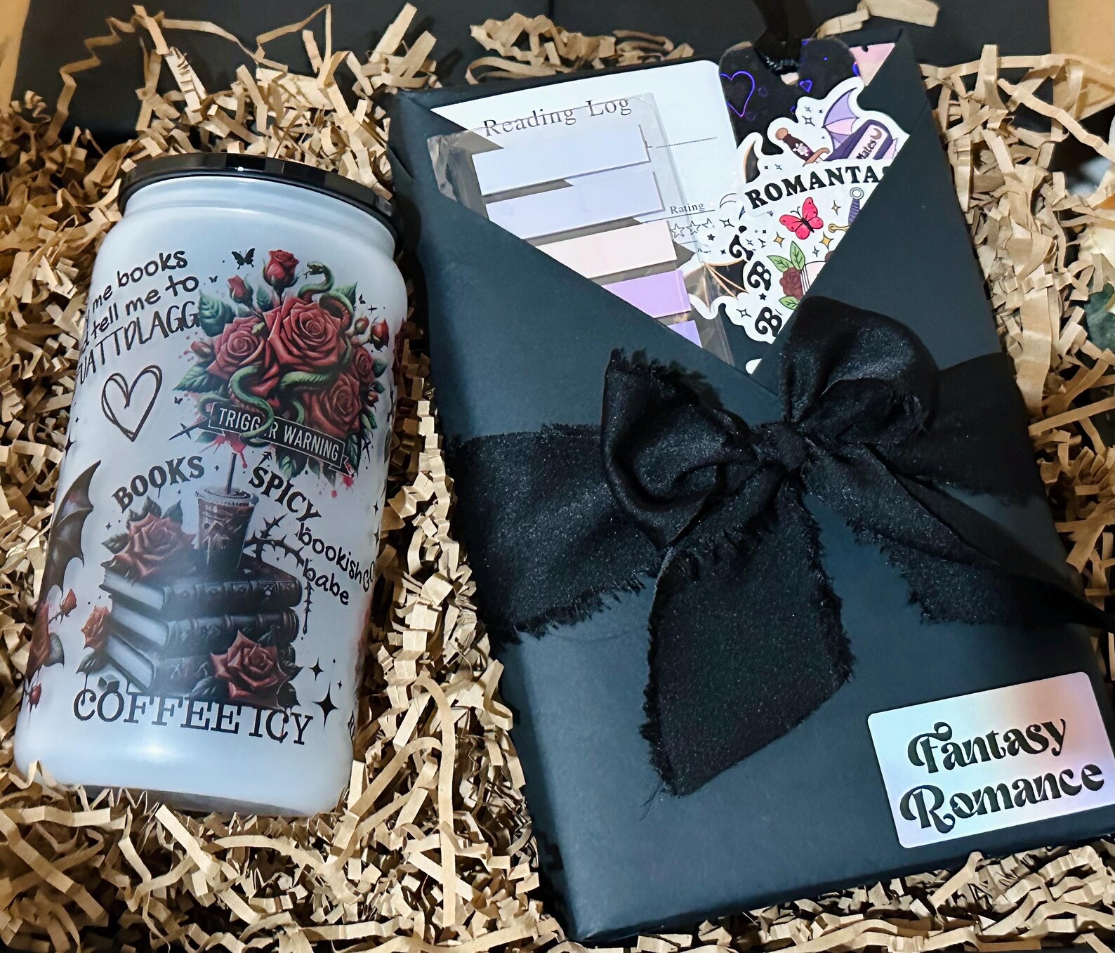 A gift set with a white tumbler featuring red roses and black gift box labeled "Fantasy Romance" with bookmarks and a reading log, surrounded by shredded paper.