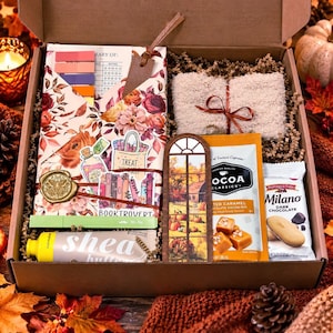 May include: A gift box containing a floral patterned journal with a leather bookmark, a pair of fuzzy beige socks, a packet of Cocoa Classics salted caramel cocoa mix, a package of Milano dark chocolate cookies, and a tube of shea butter.