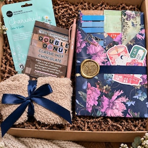 May include: A gift box containing a light blue chamomile facial mask, a packet of Double Donut classic hot chocolate, a pink pen, a beige fuzzy item with a navy blue ribbon, and a floral-patterned folder with a gold wax seal.