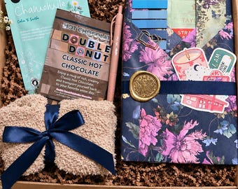 May include: A gift box containing a light blue chamomile facial mask, a packet of Double Donut classic hot chocolate, a pink pen, a beige fuzzy item with a navy blue ribbon, and a floral-patterned folder with a gold wax seal.