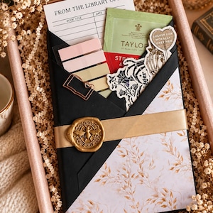 May include: A gift box containing a book wrapped in black and floral paper, secured with a gold ribbon and a bee wax seal. It includes a library checkout card, pastel sticky notes, a tea bag, and stickers featuring skeletons and text.