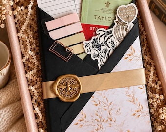 May include: A gift box containing a book wrapped in black and floral paper, secured with a gold ribbon and a bee wax seal. It includes a library checkout card, pastel sticky notes, a tea bag, and stickers featuring skeletons and text.