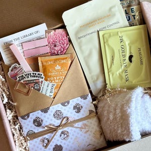 May include: A gift box filled with self-care items. Includes a sheet mask, eye mask, tea, a small white washcloth, and a decorative envelope with a bow. The envelope contains a bookmark, stickers, and a lip gloss. The box is lined with paper shreds.