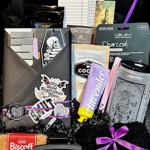May include: A gift box containing a black envelope with a skull sticker, a charcoal facial mask, a packet of cocoa, a lavender hand cream, a frosted glass tumbler with a rose graphic, a Lotus Biscoff cookie, a black hair clip, and a library card.