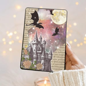 以下が含まれることがあります： 黒とグレーのケース。ファンタジーの城のデザイン、ドラゴン、満月、花のディテールが特徴です。デザインには、本のページからのテキストが含まれています。ケースは手で持たれています。