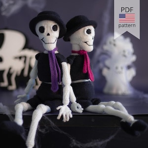 Op de afbeelding: Twee gehaakte skeletpoppen met zwarte hoeden, shirts en shorts, met witte ledematen. Eén pop heeft een paarse sjaal, de andere een roze sjaal. De achtergrond bevat Halloween-thema decoraties en de tekst "PDF pattern".