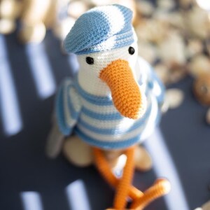 May include: A crocheted seagull toy, dressed in a blue and white striped top and a matching beret. It has an orange beak and feet, and black eyes. A charming, handmade decorative item.
